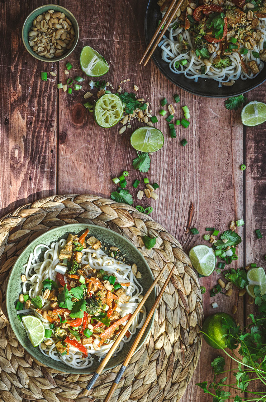 pad thai dans un bol avec des baguettes chinoises sur un sous plat en osier. Des citrons verts et des herbes décorent la table en bois